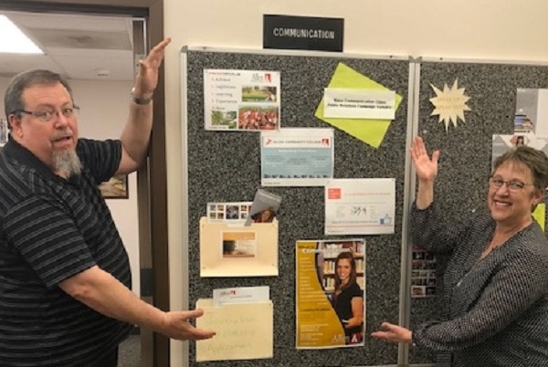 Bruce Symes and Terri Piazza try out their best Vanna White to display the Communications bulletin board in the Technology Building.